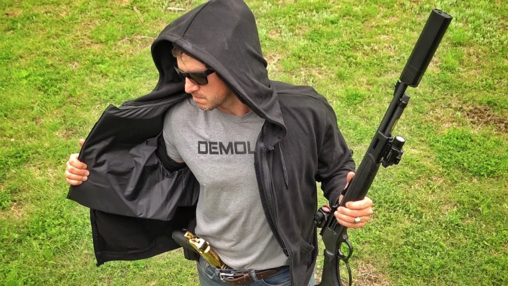 Man demonstrating concealed ballistic protection by opening black hoodie to reveal body armor underneath while holding rifle, showcasing how protective gear can be hidden in everyday clothing