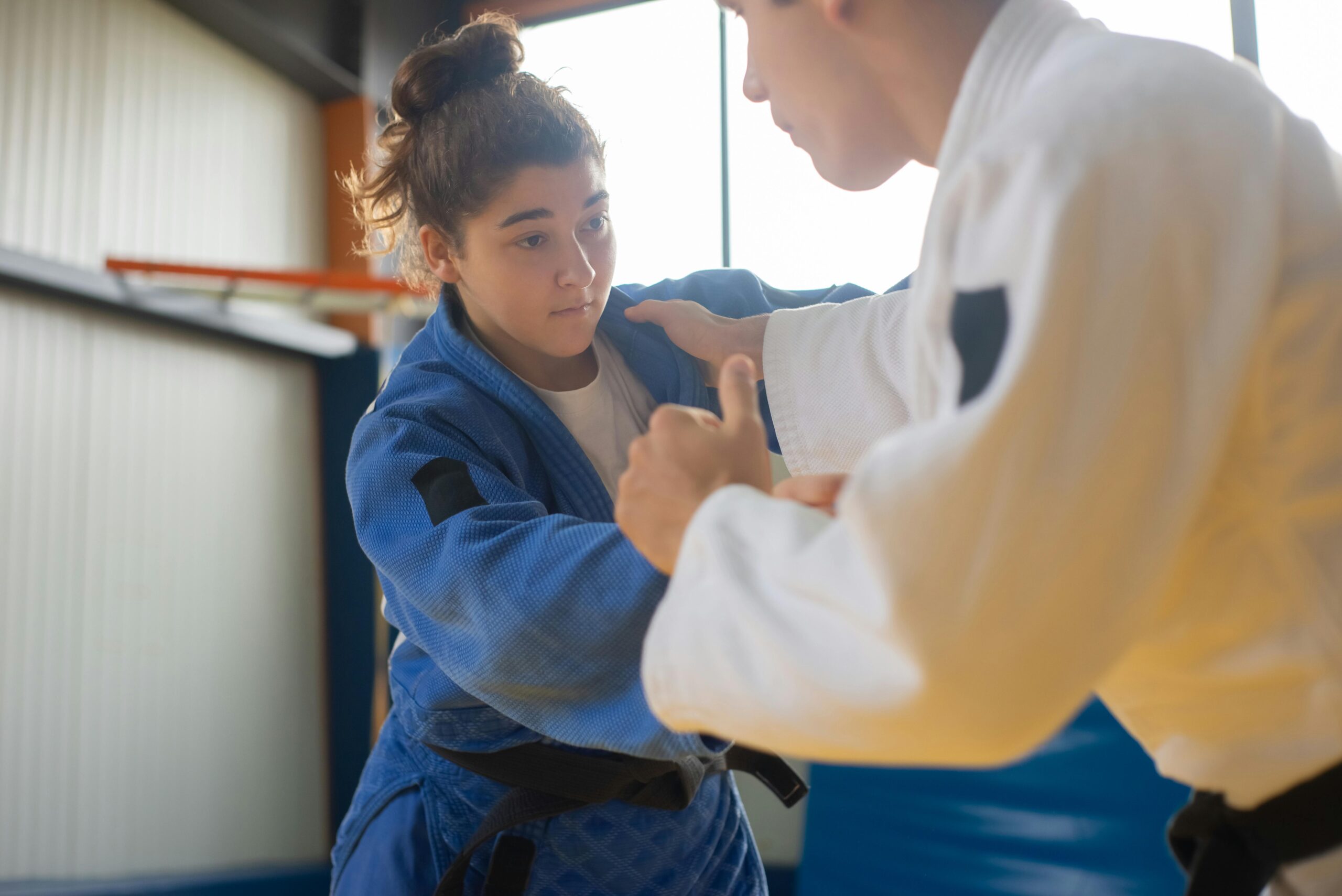 A female martial arts student in a blue gi practices a technique with a male instructor in a white gi, demonstrating the principle of passive resistance by absorbing and redirecting force.
